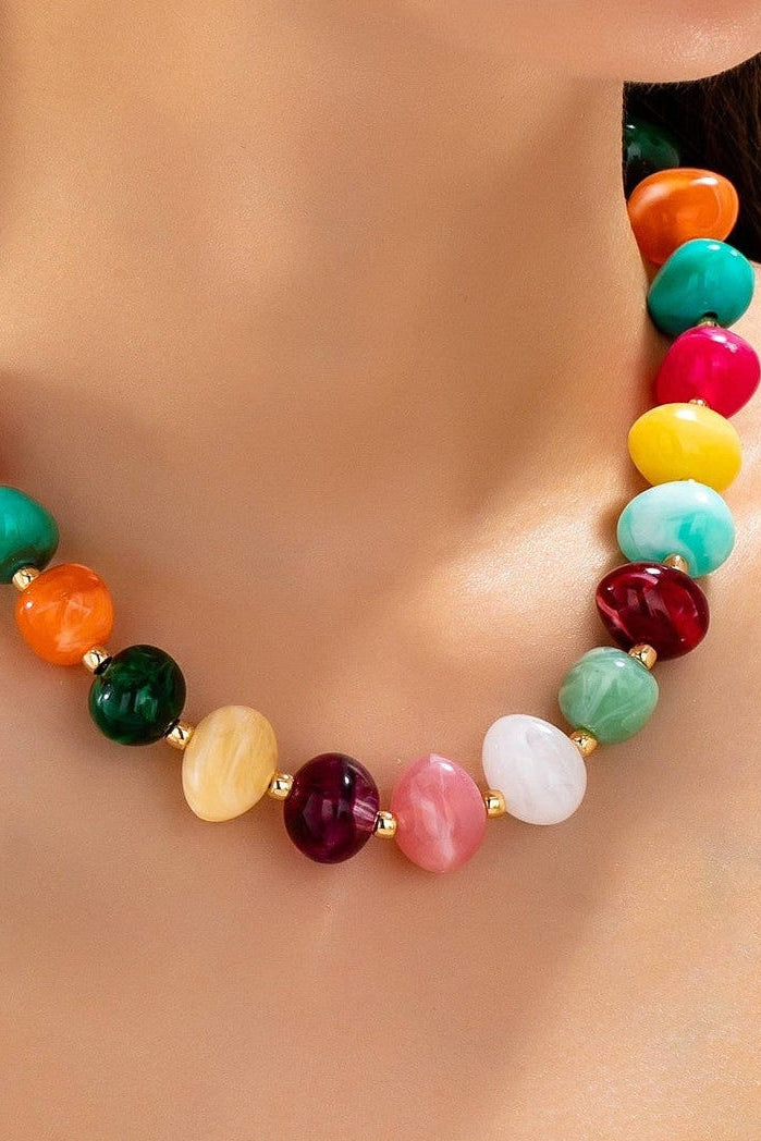 Colorful beaded statement necklace with multicolored pebble-shaped beads and gold-tone spacers, worn with a white top.