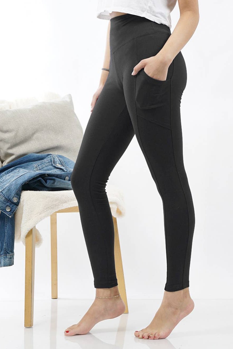 A model wearing black high-waisted leggings with side pockets, standing barefoot in a bright studio setting next to a wooden chair with a denim jacket and pillow.
