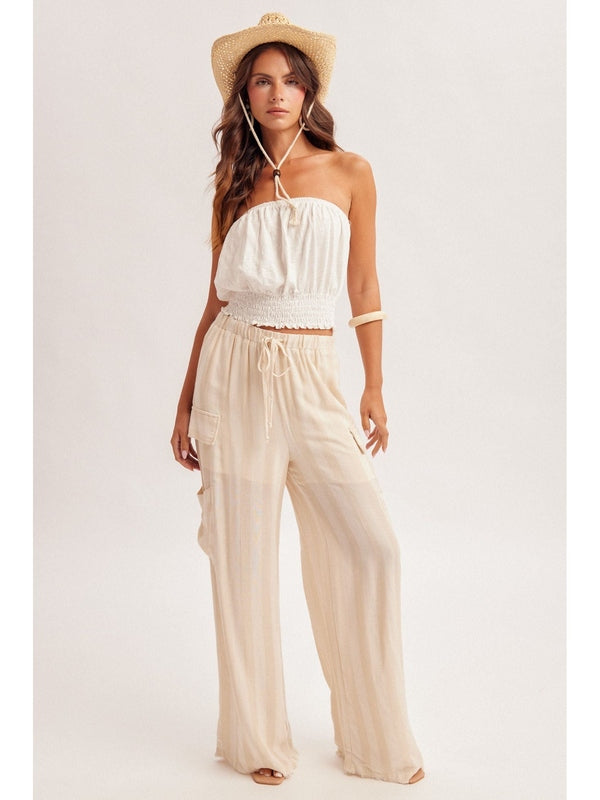 Woman wearing a white strapless top and beige wide-leg pants with a straw hat on a light gray background