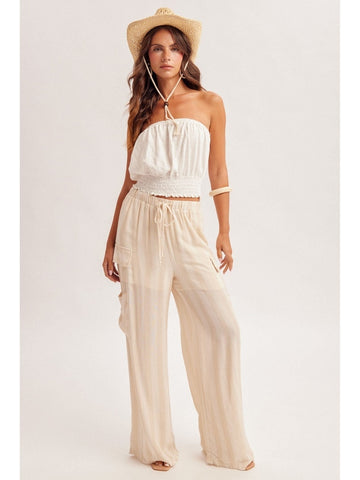 Woman wearing a white strapless top and beige wide-leg pants with a straw hat on a light gray background