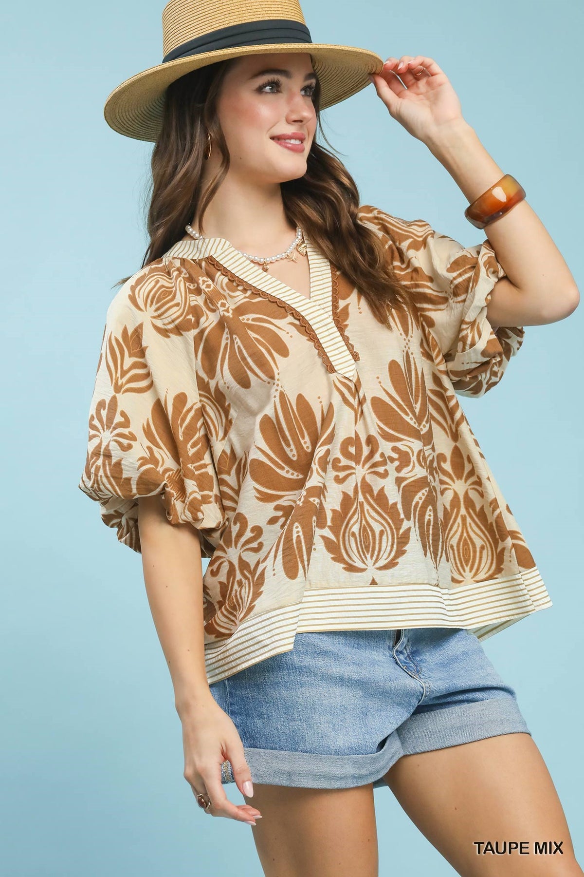 Woman wearing a beige floral blouse, denim shorts, and a straw hat on a light blue background.