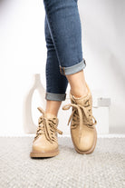A person wearing beige lace-up ankle boots with cuffed blue jeans, standing on a light textured rug against a white studio background.