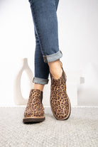 A person wearing leopard-print ankle boots with cuffed blue jeans, standing on a light textured rug against a white background.