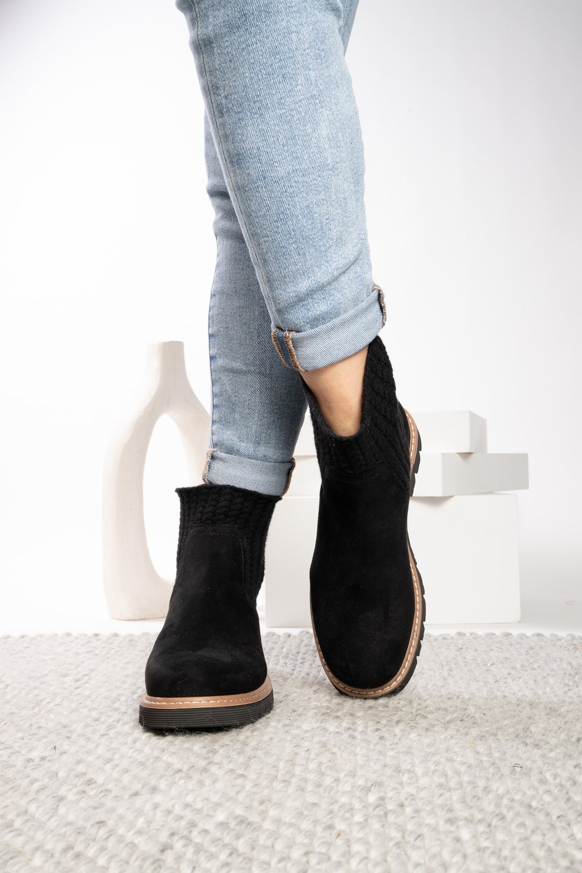A person wearing light blue cuffed jeans and black ankle boots with a knit texture and brown soles, standing on a white textured rug.
