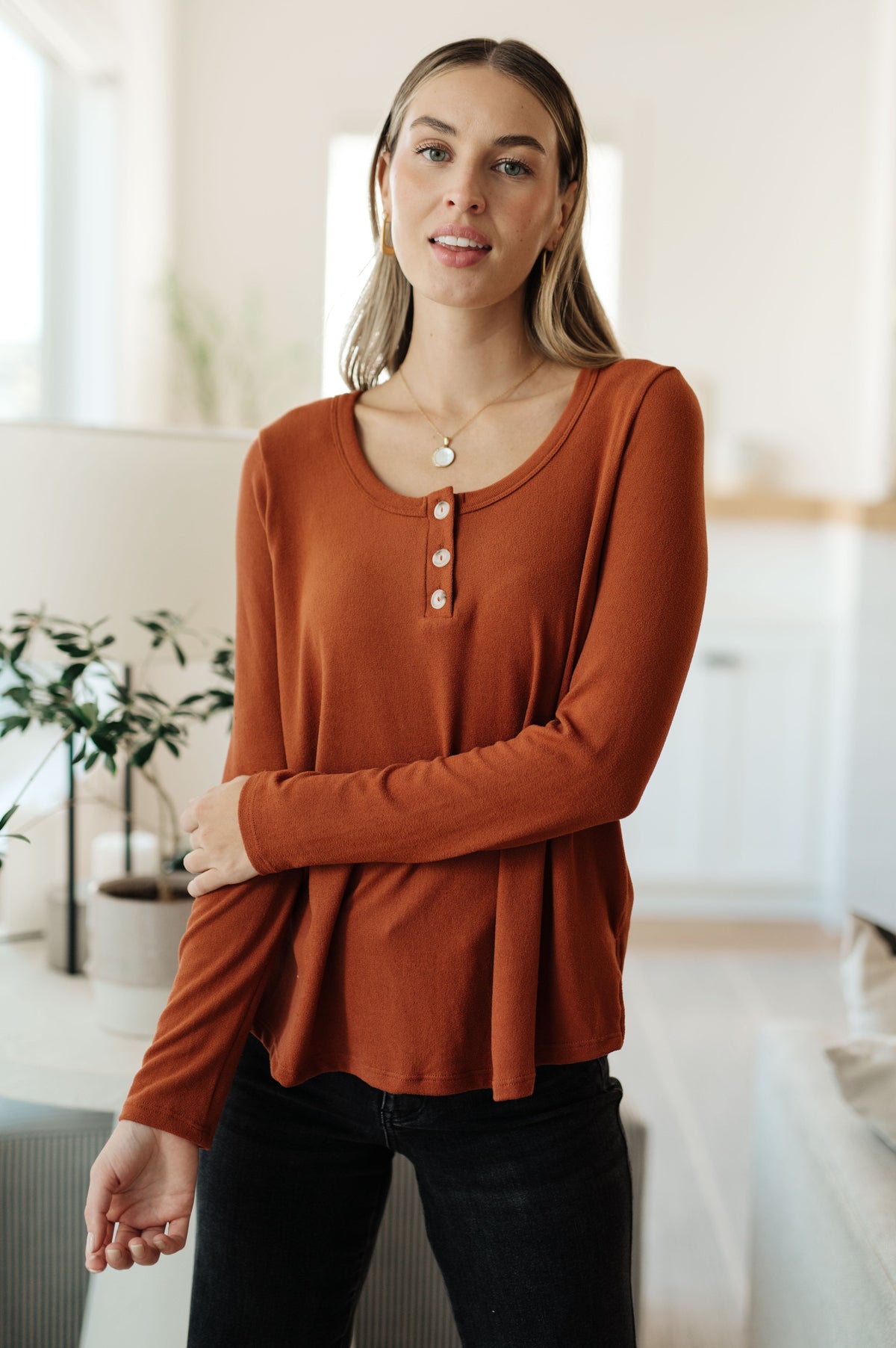 A woman modeling a rust-orange long-sleeve henley top with a round neckline and three decorative buttons, paired with black jeans in a bright, modern indoor setting.