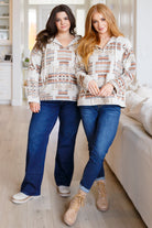 Two women modeling matching hooded sweatshirts with a Southwestern geometric pattern in beige, brown, and white, paired with blue jeans and tan boots in a bright living room.