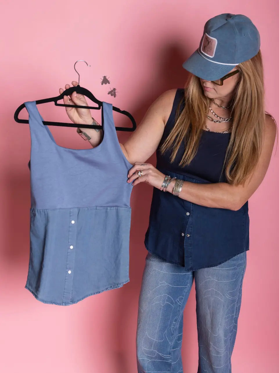 A woman wearing a blue cap and dark blue top stands against a pink wall, holding up a light blue sleeveless top on a black hanger to show it off.