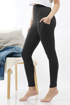 A model wearing black high-waisted leggings with side pockets, standing barefoot in a bright studio setting next to a wooden chair with a denim jacket and pillow.