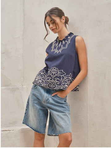Woman wearing a navy sleeveless top with white embroidery and denim shorts against a light stone wall.
