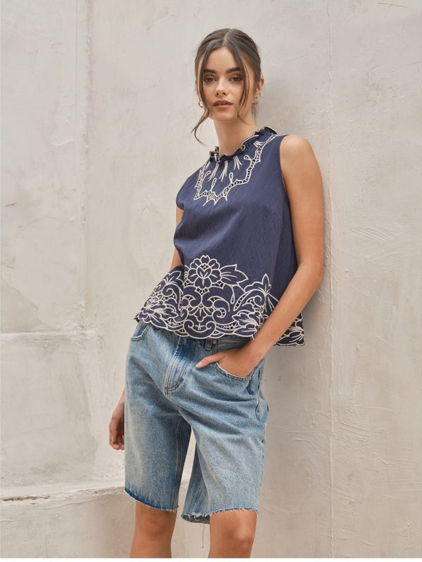 Woman wearing a navy sleeveless top with white embroidery and denim shorts against a light stone wall.