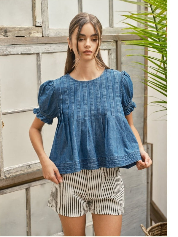 Woman wearing a blue top and striped shorts standing indoors with a plant in the background